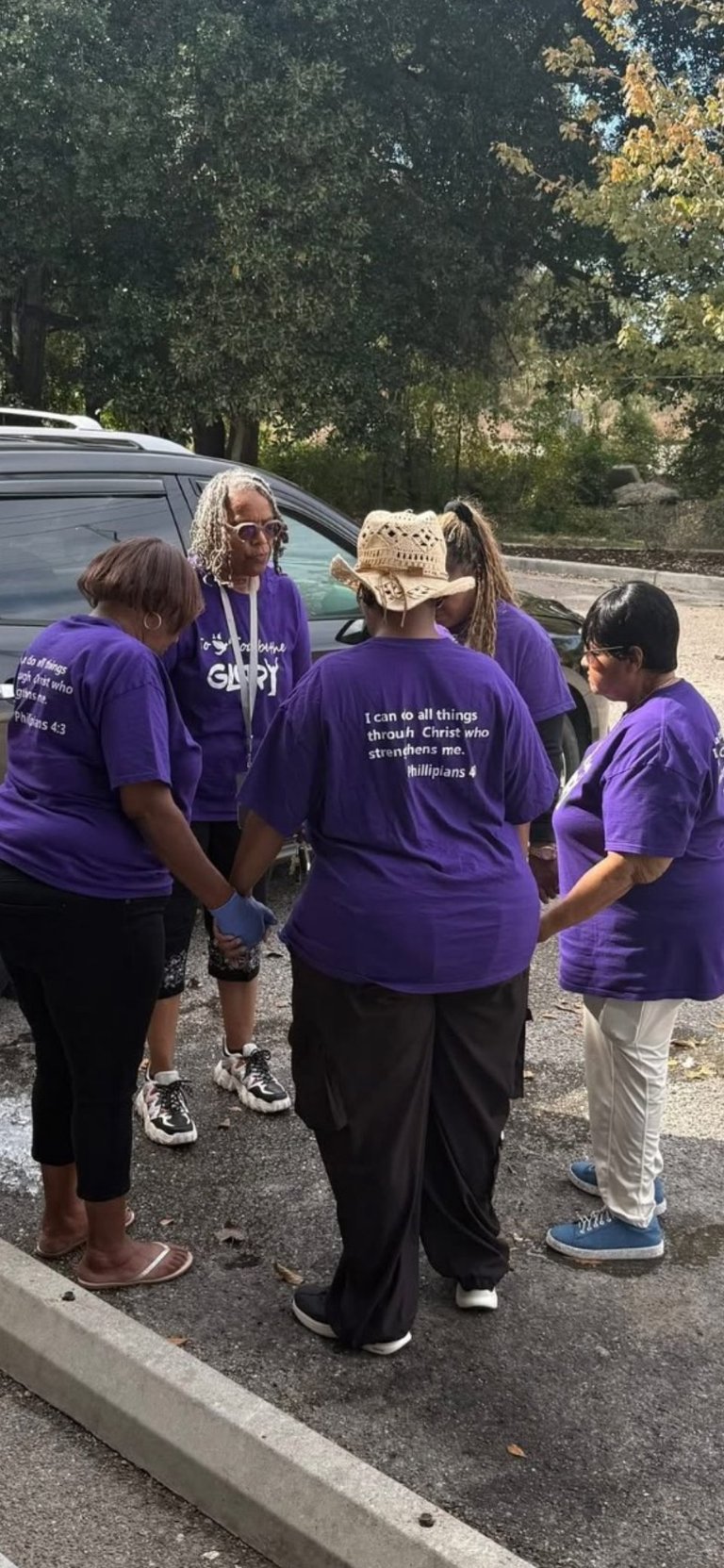 Group of four people wearing purple religious t-shirts standing in a circle in a parking lot with trees in the background