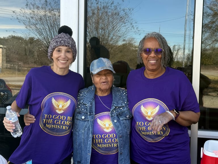 Three people wearing matching purple shirts with religious logos stand together outdoors, smiling at the camera.
